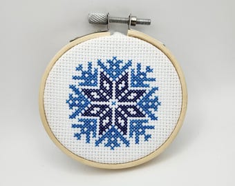 May include: A cross-stitch embroidery hoop featuring a blue and white snowflake design. The snowflake is centered on white fabric within a wooden hoop. The design uses various shades of blue to create depth and detail. The hoop has a metal tightening mechanism.