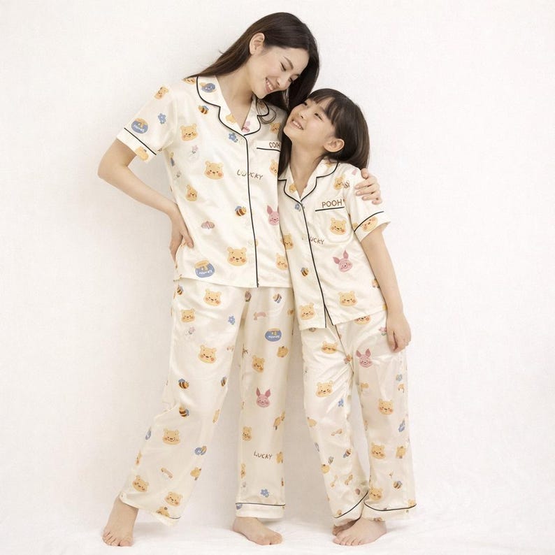 May include: Matching cream-colored pajama sets with a cartoon bear and bee pattern. The sets include short-sleeved button-up shirts and pants, trimmed with black piping. The word "Lucky" is printed on the pants.