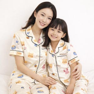 May include: Matching cream-colored pajama sets featuring Winnie the Pooh characters. The sets include a button-down shirt and pants, with black trim and the word "Lucky" printed on the shirt. The shirt of the child's set says "Pooh".