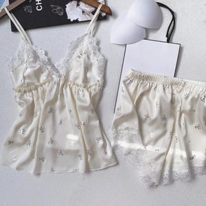 May include: Ivory satin pajama set with a camisole top and shorts. The top and shorts are adorned with silver bow accents and lace trim. The camisole has delicate spaghetti straps and a V-neckline. A white bra and shopping bag are also in the image.