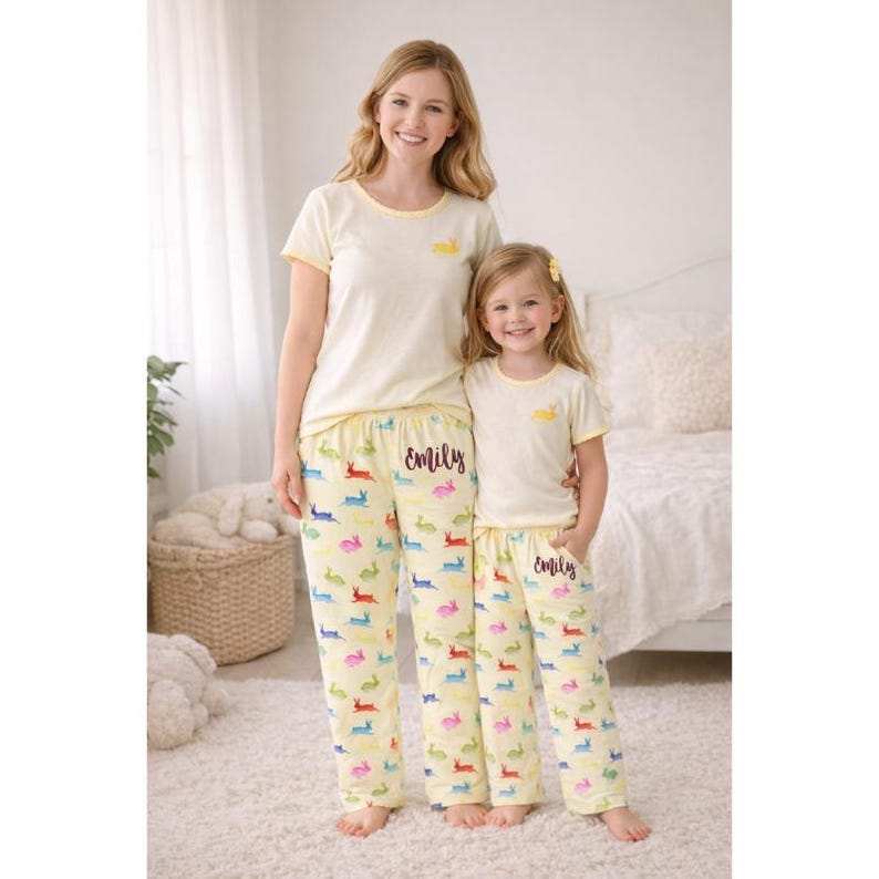 Puede incluir: Conjuntos de pijama color crema a juego para madre e hija. Las camisetas tienen ribetes amarillos y un peque&ntilde;o gr&aacute;fico de conejito amarillo. Los pantalones de pijama son de color amarillo claro con estampados de conejitos coloridos y el nombre "Emily" en escritura negra.