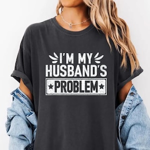May include: Dark gray t-shirt with the text "I'M MY HUSBAND'S PROBLEM" in white. The text is arranged in a stacked format with a decorative border around the word "PROBLEM". The shirt has rolled-up sleeves and is worn with a denim jacket.