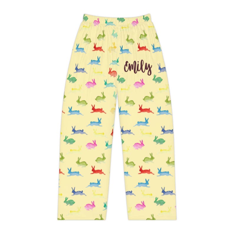 Puede incluir: Pantalones de pijama amarillos claros con un patr&oacute;n repetido de conejos coloridos en verde, azul, rojo y rosa. El nombre "emily" est&aacute; escrito en cursiva en los pantalones.