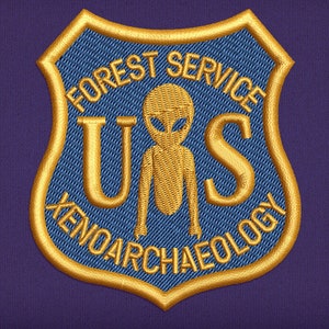 May include: Embroidered patch featuring a shield design with the text "FOREST SERVICE" and "XENOARCHAEOLOGY". The patch includes a gold alien figure and the letters "U S". The patch is blue with gold and white stitching.