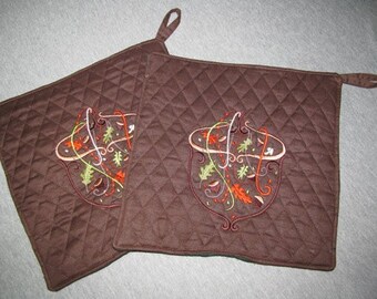 May include: Two brown quilted pot holders with a fall-themed embroidered design of a basket filled with leaves and acorns.