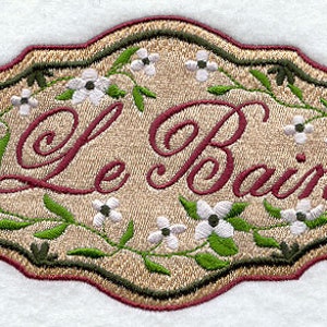 May include: A beige embroidered oval sign with a green floral border and the text "Le Bain" in red cursive lettering.