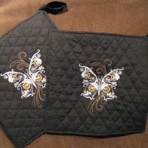 May include: Two black quilted pot holders featuring a butterfly design. The butterfly is embroidered with white, gold, and silver details, resembling gears and mechanical elements. Each pot holder has a black loop for hanging.