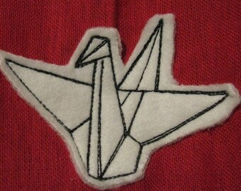 May include: White felt origami crane with black stitching, on a red fabric background.