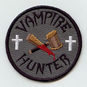 May include: A gray circular embroidered patch with the words "Vampire Hunter" in a white outline around the edge. A crossed wooden stake and hammer with a red blood drip are in the center. Two white crosses are on either side of the stake and hammer.