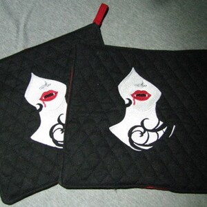 May include: Two black quilted pot holders with a vampire-themed design. Each pot holder features an embroidered image of a woman's face with white skin, black hair, and red lips with fangs. Red loops are attached for hanging.