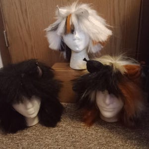 Puede incluir: Tres cabezas de maniquí blancas con pelucas de piel sintética con orejas de animal. Una peluca es negra sólida, otra es blanca con manchas naranjas y negras, y otra es una mezcla de negro, naranja y blanco. Cada peluca tiene orejas puntiagudas.