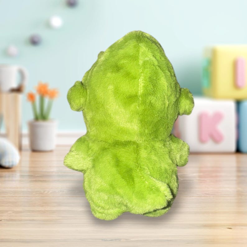 May include: A lime green plush toy, viewed from the back, with a hood and small ear-like protrusions. The toy has a soft, fuzzy texture and is likely designed for children. The background is blurred, suggesting a nursery or playroom setting.