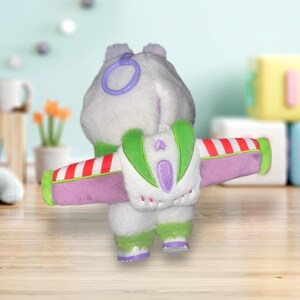 May include: A plush toy in the shape of a character from a popular animated film, viewed from the back. The toy is primarily white with green, purple, and red accents. It has a backpack with wings and a purple ring attached to its head.