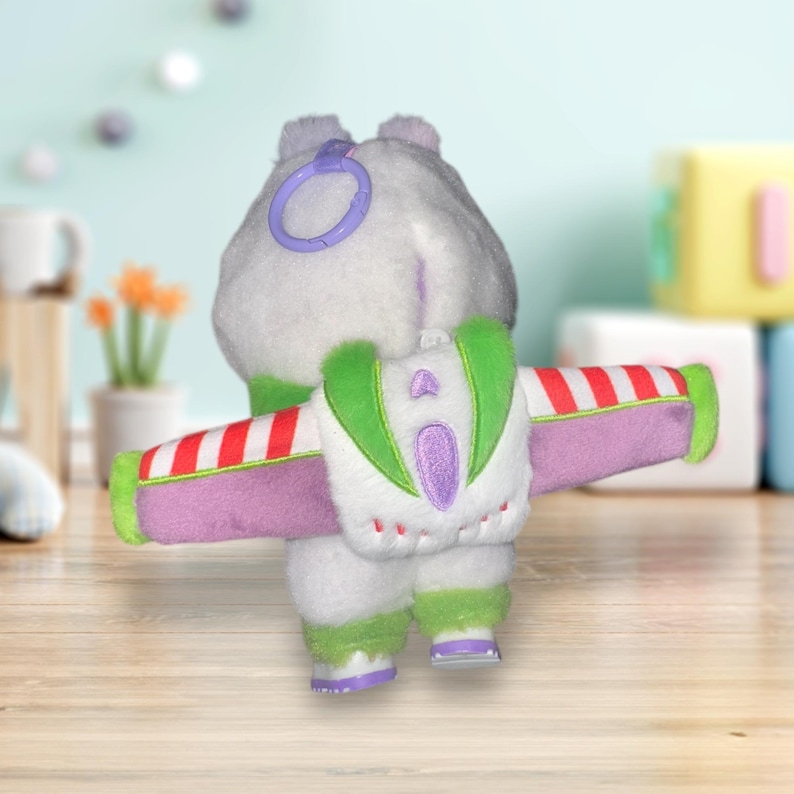 May include: A plush toy in the shape of a character from a popular animated film, viewed from the back. The toy is primarily white with green, purple, and red accents. It has a backpack with wings and a purple ring attached to its head.