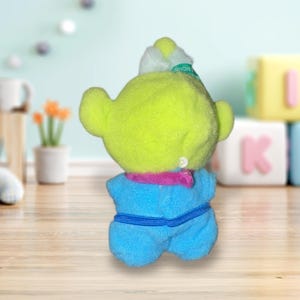 May include: A plush toy alien with a lime green head, large ears, and a blue outfit. The alien has a pink scarf and a white hat with the word "SNOW" on it. The toy is sitting on a wooden surface.