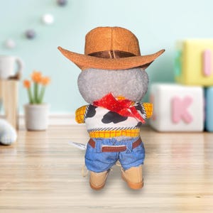 May include: A plush toy dressed as a cowboy, viewed from the back. It wears a brown cowboy hat, a cow-print vest, blue denim shorts with a brown belt, a red neckerchief, and tan boots. The toy is on a wooden surface.