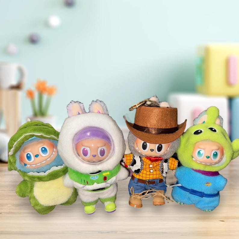 May include: Four plush dolls in character costumes. A green dinosaur, a white space suit, a cowboy, and a green alien. The dolls have large eyes and are on a light brown wooden surface.