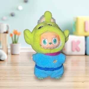 May include: A plush toy featuring a character in a green alien costume with a blue jumpsuit. The toy has a smiling face with large, round eyes and a small mouth. The costume has a hood and a belt. The toy is designed for children.