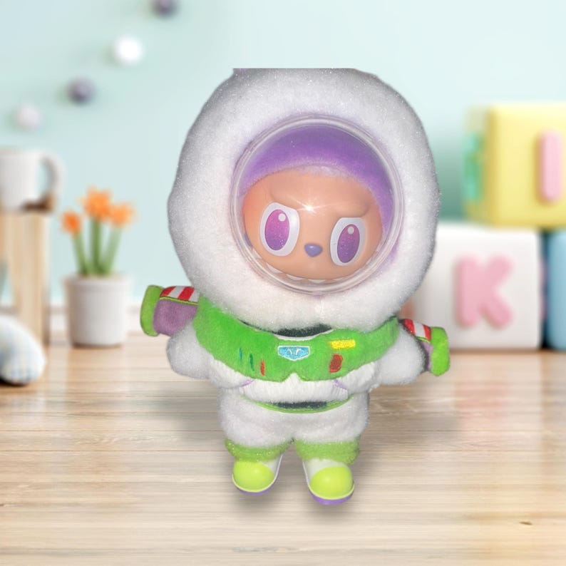 May include: A plush toy of a character in a white spacesuit with green and purple accents. The toy has a white hood with a clear visor, and the character's face is visible. The toy is approximately 6 inches tall.