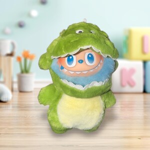 May include: A plush toy in the shape of a green dinosaur with a yellow belly. The toy has a smiling face with blue eyes and a blue furry collar. The toy is designed to look like a dinosaur is eating a smiling face.