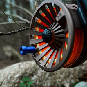 K&ouml;nnte beinhalten: Nahaufnahme einer Fliegenfischrolle mit grauem Rahmen und orangefarbener Schnur. Die Rolle hat einen blauen Griff und befindet sich vor einem verschwommenen nat&uuml;rlichen Hintergrund aus B&auml;umen und Felsen. Die Rolle ist an einer schwarzen Rute befestigt.
