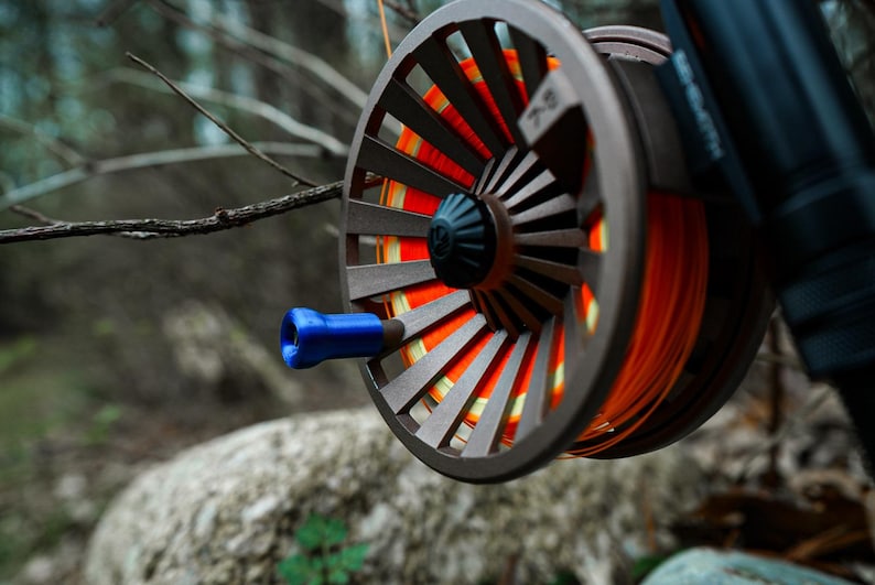 K&ouml;nnte beinhalten: Nahaufnahme einer Fliegenfischrolle mit grauem Rahmen und orangefarbener Schnur. Die Rolle hat einen blauen Griff und befindet sich vor einem verschwommenen nat&uuml;rlichen Hintergrund aus B&auml;umen und Felsen. Die Rolle ist an einer schwarzen Rute befestigt.