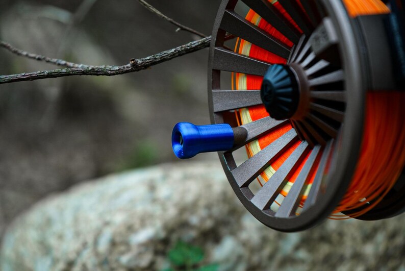 K&ouml;nnte beinhalten: Nahaufnahme einer Fliegenfischrolle mit braunem Rahmen und blauem Griff. Die Rolle hat orangefarbene und gelbe Angelschnur. Der Hintergrund ist unscharf.