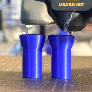 K&ouml;nnte beinhalten: Zwei blaue, zylindrische Objekte werden im 3D-Druckverfahren hergestellt. Die Objekte haben eine ausgestellte Oberseite und eine geriffelte Textur. Der Druckkopf und die Worte "CAUTION HOT" sind im Hintergrund zu sehen.