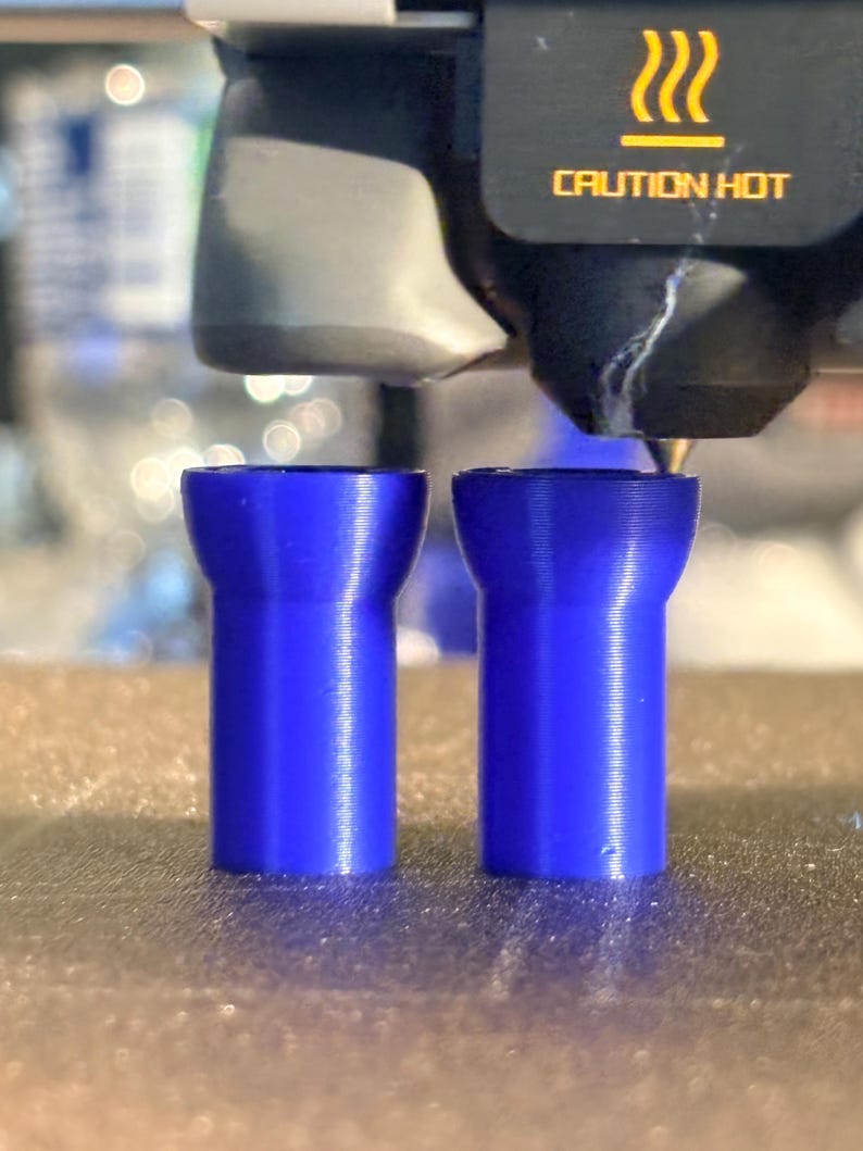 K&ouml;nnte beinhalten: Zwei blaue, zylindrische Objekte werden im 3D-Druckverfahren hergestellt. Die Objekte haben eine ausgestellte Oberseite und eine geriffelte Textur. Der Druckkopf und die Worte "CAUTION HOT" sind im Hintergrund zu sehen.