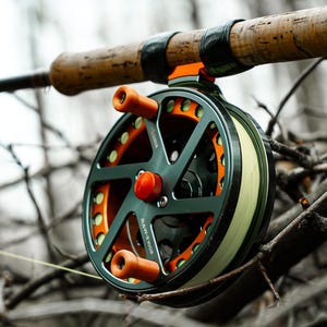 K&ouml;nnte beinhalten: Eine Fliegenfischrolle mit dunkelgr&uuml;nem Rahmen und orangefarbenen Akzenten ist an einer Rute mit Korkgriff befestigt. Die Rolle hat einen leuchtend orangefarbenen Mittelknopf und orangefarbene Griffe. Der Hintergrund ist verschwommen und zeigt &Auml;ste.