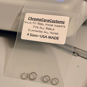 K&ouml;nnte beinhalten: Eine transparente Plastikt&uuml;te mit kleinen, silberfarbenen Rollenknauf-Eins&auml;tzen. Das Etikett lautet "ChromeCoreCustoms" und listet Funktionen wie "Snug Fit", "Fits All Reels" und "Eliminates All Noise" auf. Auf dem Etikett steht auch "4 Sizes USA MADE".