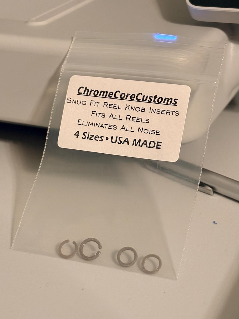 K&ouml;nnte beinhalten: Eine transparente Plastikt&uuml;te mit kleinen, silberfarbenen Rollenknauf-Eins&auml;tzen. Das Etikett lautet "ChromeCoreCustoms" und listet Funktionen wie "Snug Fit", "Fits All Reels" und "Eliminates All Noise" auf. Auf dem Etikett steht auch "4 Sizes USA MADE".