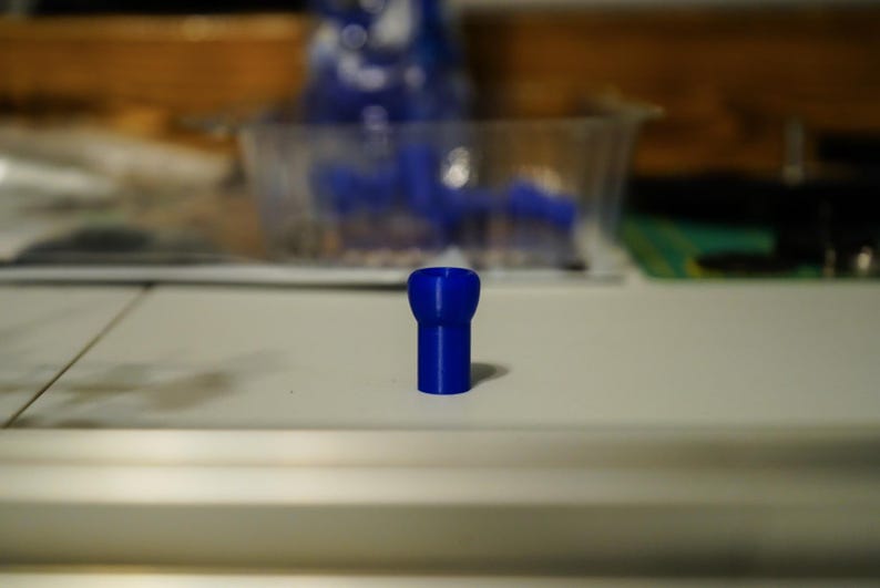 K&ouml;nnte beinhalten: Ein kleiner, leuchtend blauer Kunststoffgegenstand mit zylindrischem Sockel und breiterem Oberteil. Der Gegenstand steht auf einer wei&szlig;en Oberfl&auml;che. Weitere blaue Objekte sind im Hintergrund zu sehen.