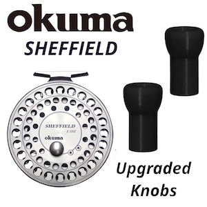 May include: A silver Okuma Sheffield S 1002 fly fishing reel with upgraded black knobs. The reel has a circular design with multiple holes and the text "SHEFFIELD" and "okuma" in black.