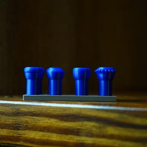 May include: Four blue, cylindrical objects with varying top designs are arranged on a gray base. The objects are set against a dark, wooden background. The items appear to be small, possibly decorative or functional.