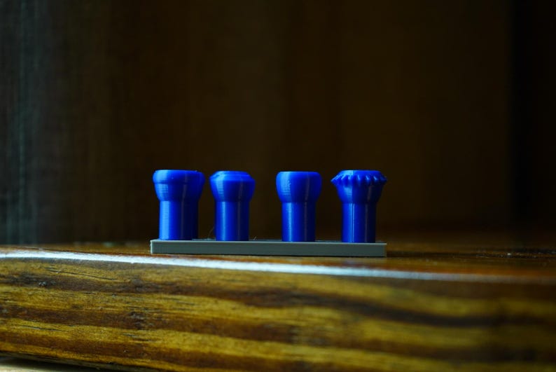 May include: Four blue, cylindrical objects with varying top designs are arranged on a gray base. The objects are set against a dark, wooden background. The items appear to be small, possibly decorative or functional.