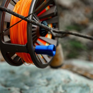 May include: Close-up of a fly fishing reel with bright orange line. The reel is dark brown with a blue handle and wooden grip. A tree branch is in the foreground, and the background is blurred.