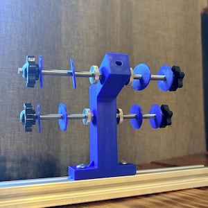 Könnte beinhalten: Ein blaues 3D-gedrucktes Gerät mit mehreren einstellbaren Komponenten. Es verfügt über eine vertikale Stützstruktur mit horizontalen Stangen, blauen Scheiben und schwarzen Knöpfen. Das Gerät ist auf einer silbernen Schiene montiert, was auf ein Präzisionswerkzeug hindeutet.