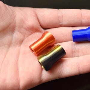 K&ouml;nnte beinhalten: Nahaufnahme von drei kleinen, zylindrischen Objekten. Zwei sind im Fokus: eines ist orange, das andere schwarz und gold. Ein blaues Objekt ist teilweise sichtbar. Alle haben eine ausgestellte Oberseite und ein Loch in der Mitte.