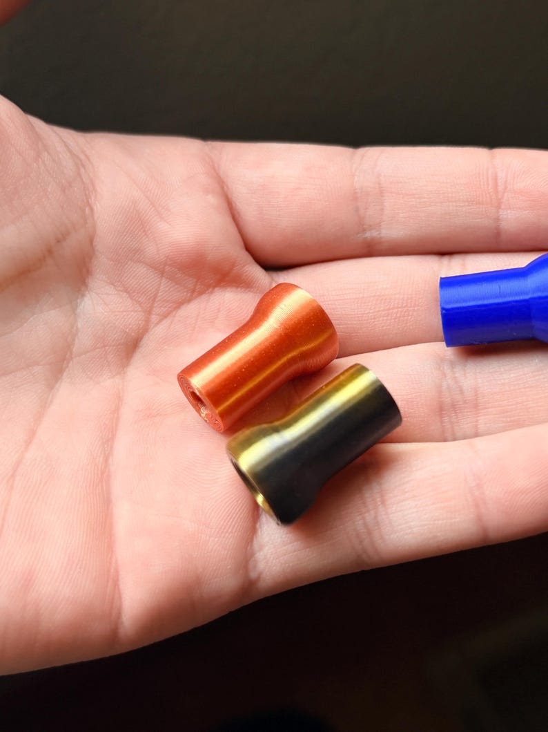 K&ouml;nnte beinhalten: Nahaufnahme von drei kleinen, zylindrischen Objekten. Zwei sind im Fokus: eines ist orange, das andere schwarz und gold. Ein blaues Objekt ist teilweise sichtbar. Alle haben eine ausgestellte Oberseite und ein Loch in der Mitte.