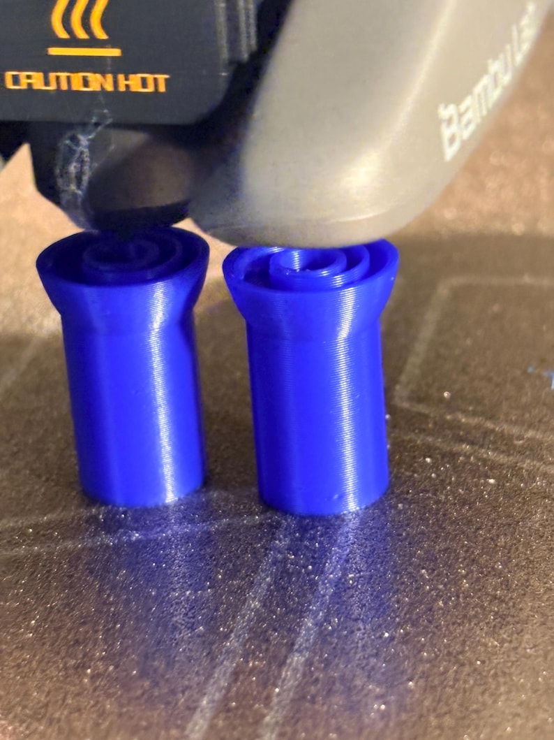 K&ouml;nnte beinhalten: Zwei leuchtend blaue, zylindrische Objekte mit einem ausgestellten oberen Teil werden von einem 3D-Drucker erstellt. Der Druckkopf ist &uuml;ber den Objekten sichtbar. Der Text "CAUTION HOT" ist auf dem Drucker aufgedruckt.