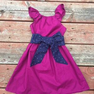 May include: A magenta dress with a navy blue polka dot bow. The dress has ruffled straps and a gathered bodice.