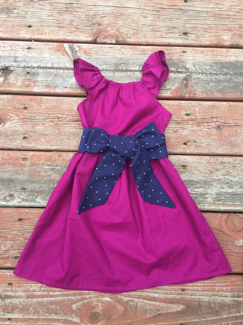 May include: A magenta dress with a navy blue polka dot bow. The dress has ruffled straps and a gathered bodice.