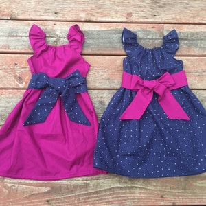 May include: Two sleeveless dresses with a bow at the waist. One dress is a solid magenta color and the other is navy blue with white polka dots. Both dresses have ruffled sleeves.