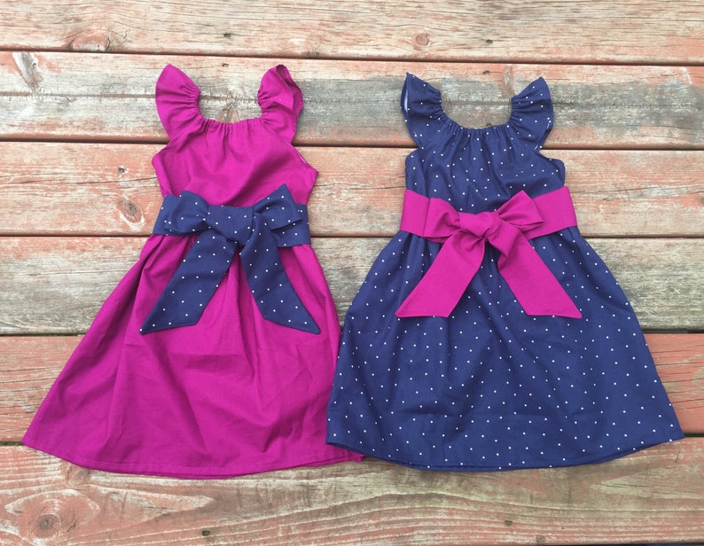 May include: Two sleeveless dresses with a bow at the waist. One dress is a solid magenta color and the other is navy blue with white polka dots. Both dresses have ruffled sleeves.