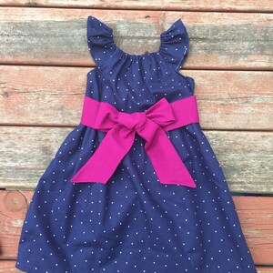 May include: A navy blue dress with white polka dots and a fuchsia satin bow. The dress has short, ruffled sleeves and a scoop neckline.