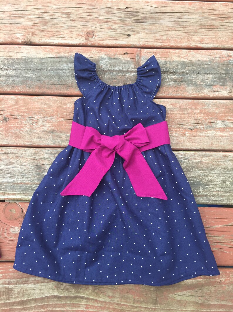 May include: A navy blue dress with white polka dots and a fuchsia satin bow. The dress has short, ruffled sleeves and a scoop neckline.