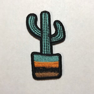 May include: A green cactus with three arms in a black pot with orange and brown stripes. The cactus is embroidered on a white background.