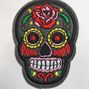May include: Embroidered gray skull patch with a red rose, yellow and red accents, and white teeth. The patch features a detailed design with colorful stitching, perfect for adding to clothing or accessories. The patch is a Day of the Dead theme.