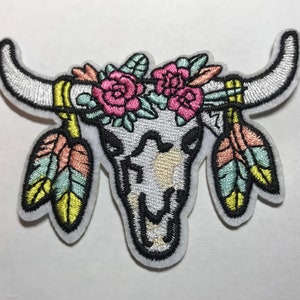 May include: White embroidered patch of a cow skull with a floral crown of pink and green flowers and two turquoise and yellow feathers on each side.
